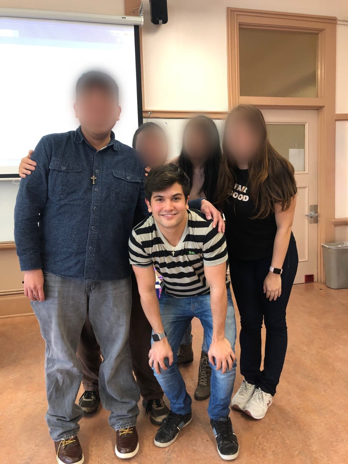 Matheus Siqueira agachado à frente de quatro pessoas de rosto desfocado, numa sala com tela de projeção e quadro branco.