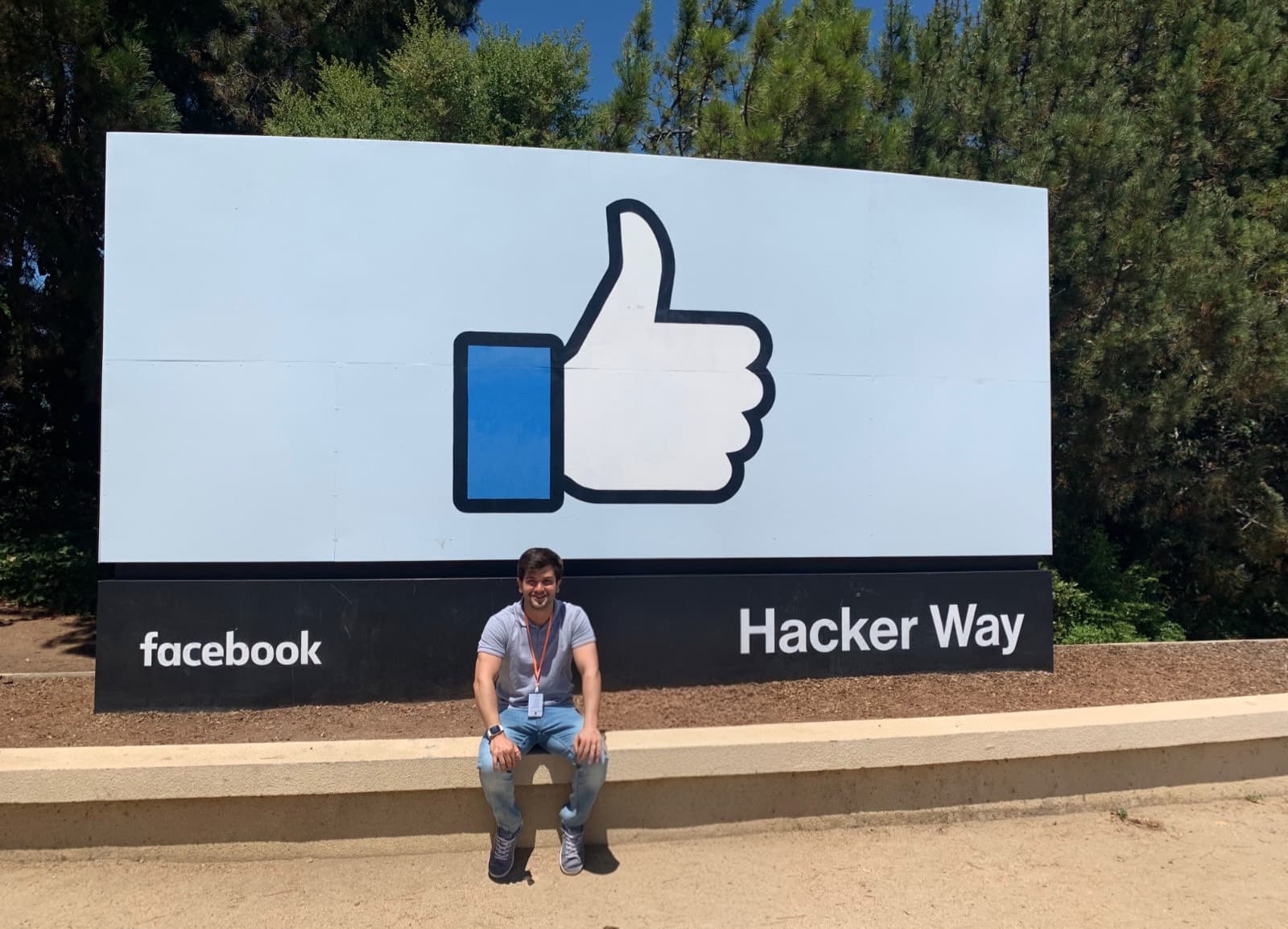 Matheus Siqueira sentado diante da placa do Facebook com o símbolo de curtir e o nome Hacker Way.